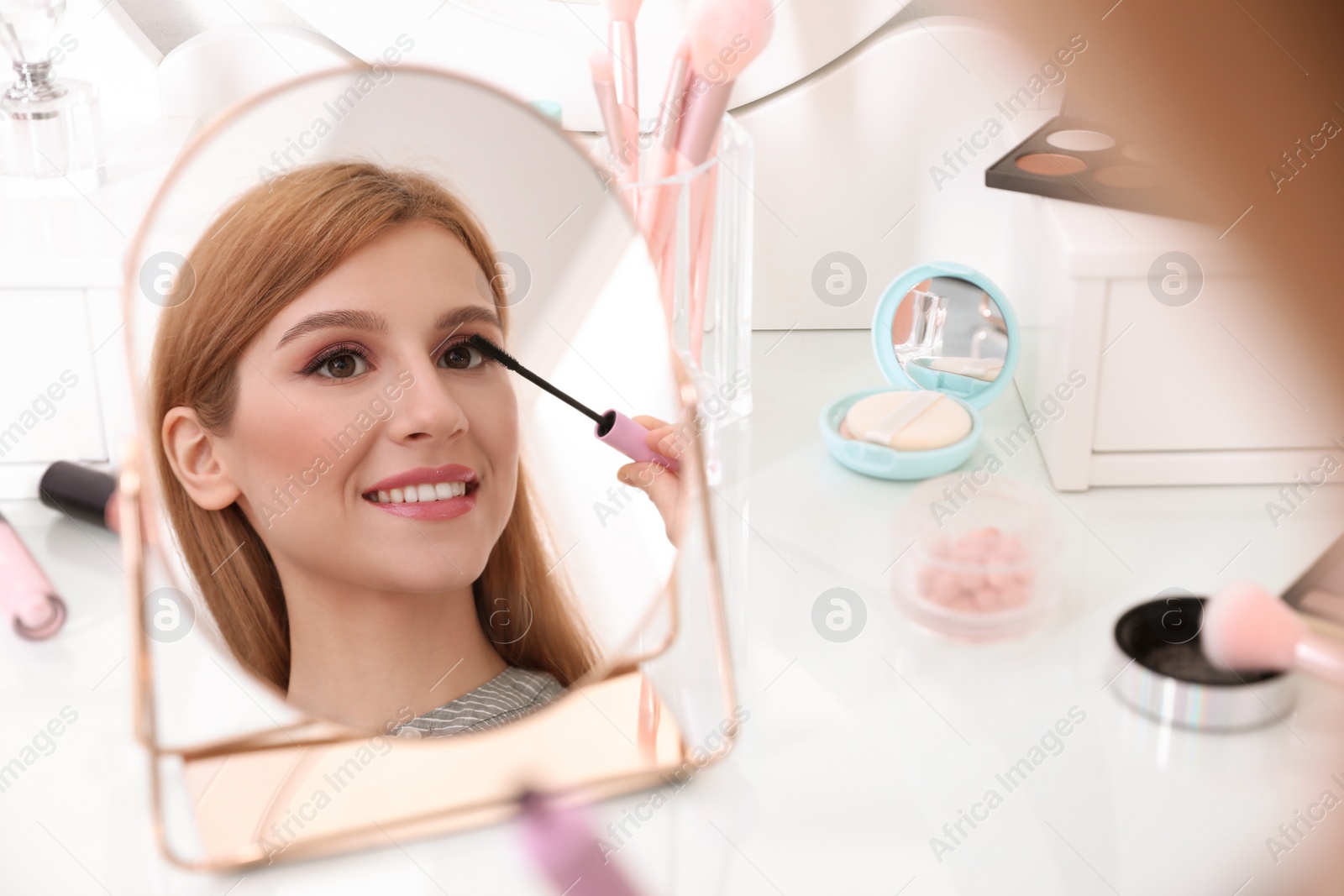 Photo of Beautiful young woman applying makeup near mirror indoors