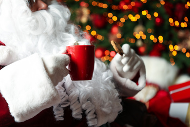 Photo of Santa Claus with hot drink and cookie against blurred Christmas lights, closeup