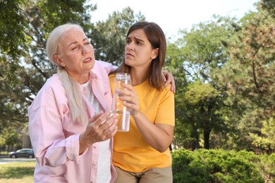 Photo of Woman with bottle of water helping mature lady in park. Suffering from heat stroke