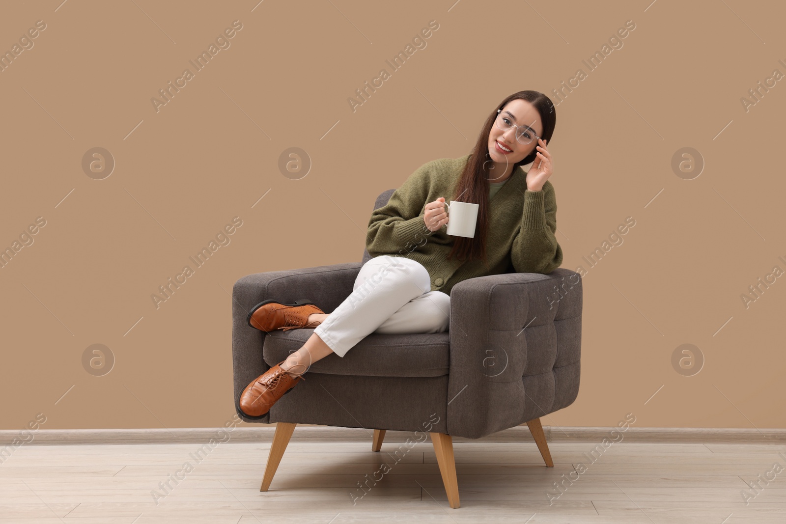 Photo of Beautiful woman with cup of drink sitting in armchair near beige wall indoors