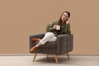 Photo of Beautiful woman with cup of drink sitting in armchair near beige wall indoors