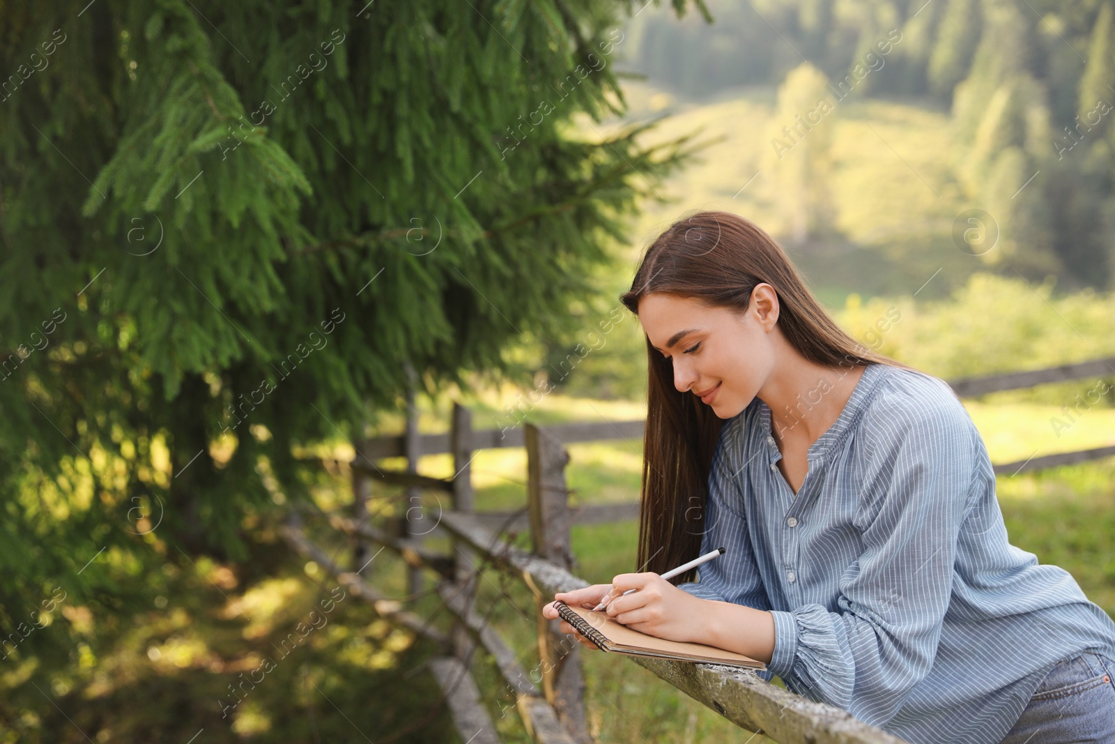 Photo of Beautiful young woman drawing with pencil in notepad near wooden fence