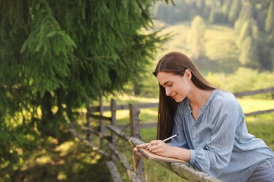 Photo of Beautiful young woman drawing with pencil in notepad near wooden fence