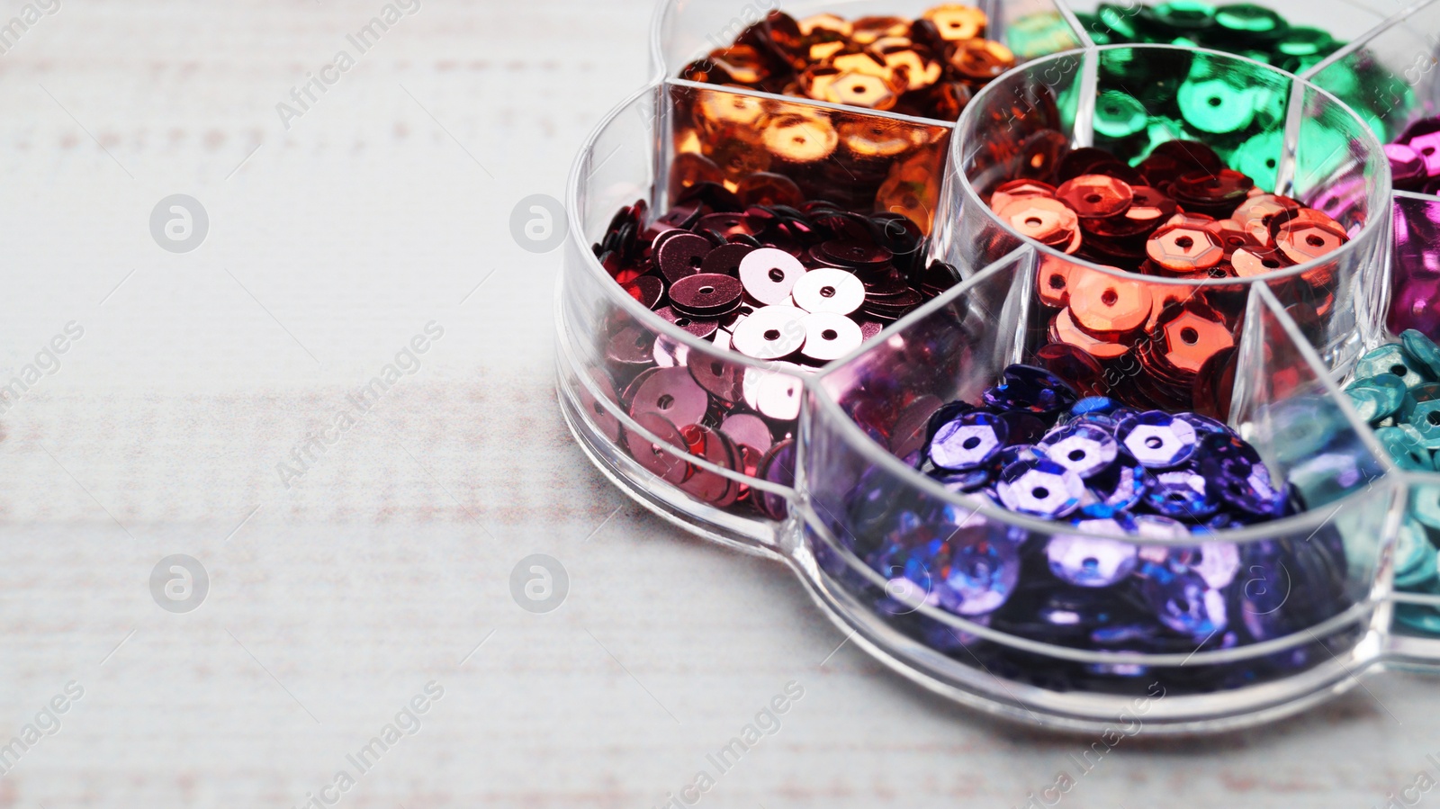 Photo of Container with many colorful sequins on white wooden table, closeup. Space for text