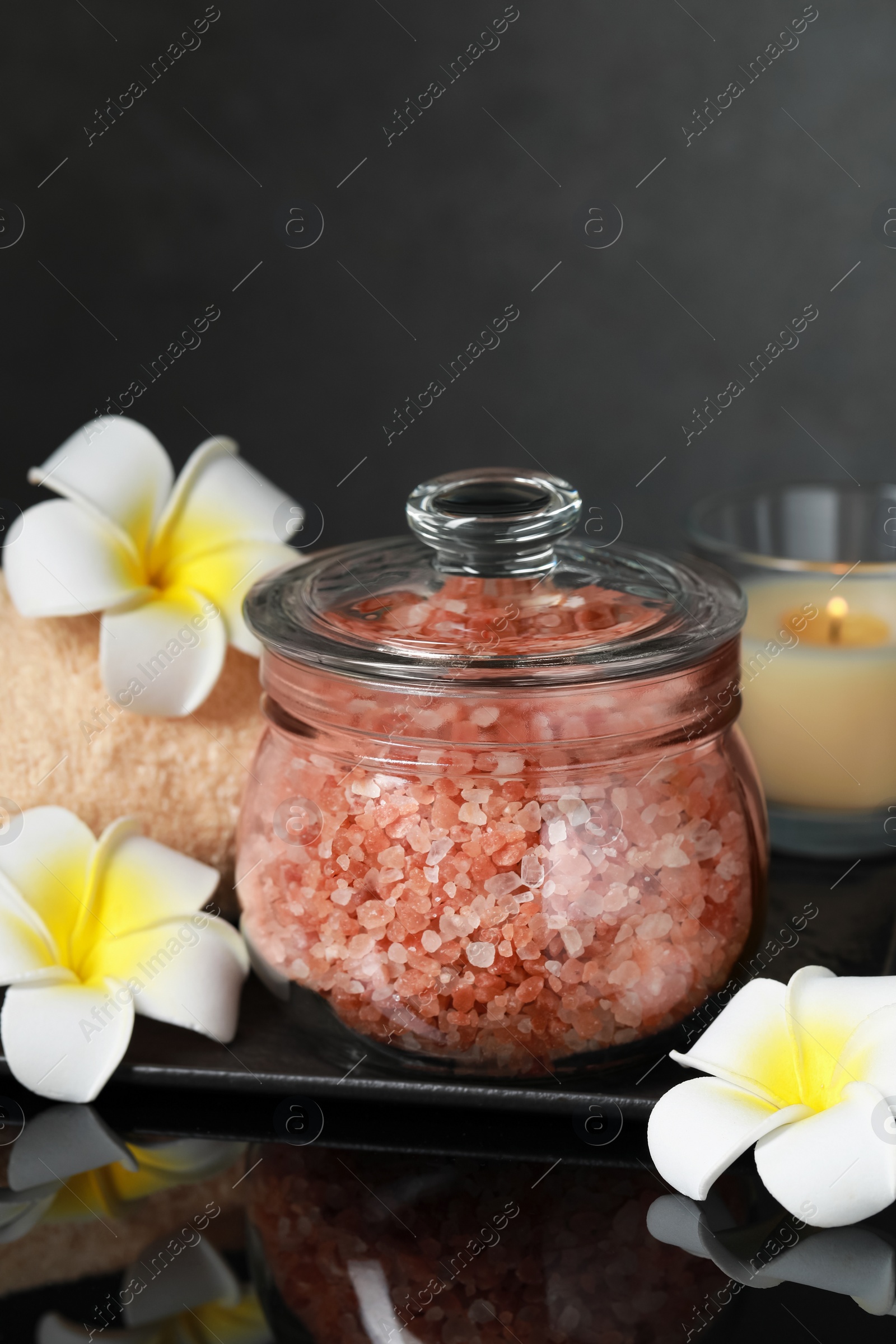 Photo of Glass jar with natural sea salt and beautiful flowers on mirror surface