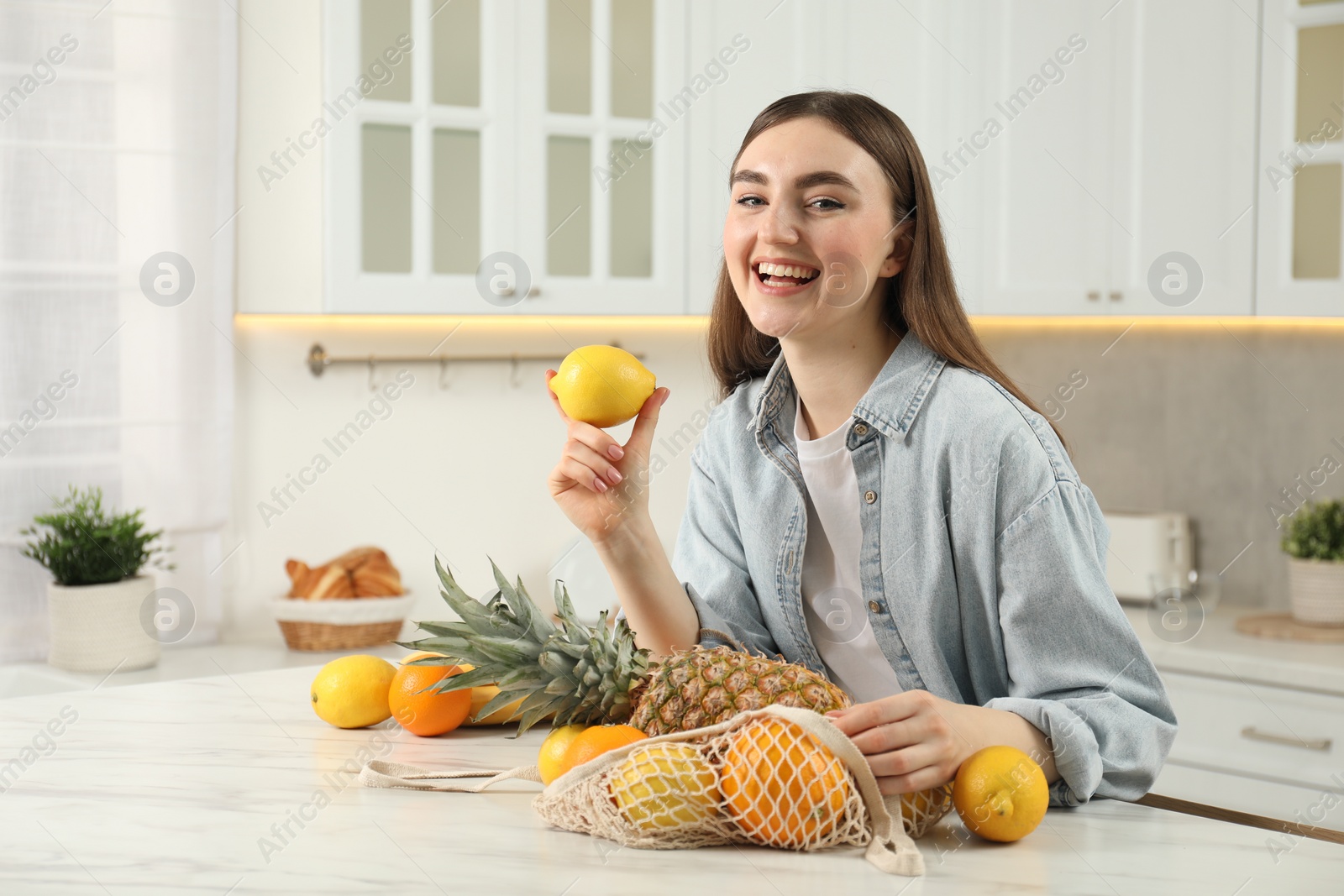 Photo of Woman with lemon and string bag of fresh fruits at light marble table in kitchen