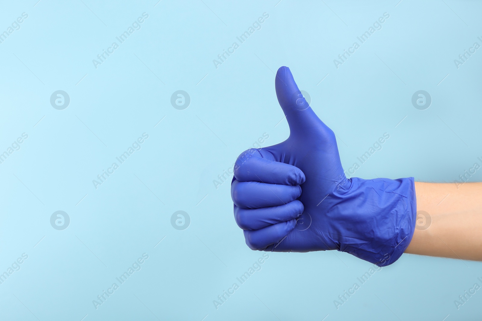 Photo of Person in medical gloves showing thumb up on light blue background, closeup of hand. Space for text