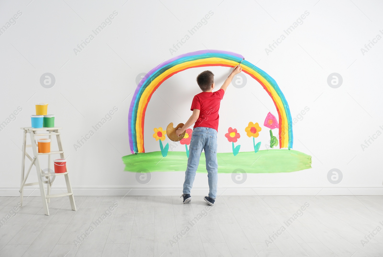 Image of Little child drawing rainbow and flowers on white wall indoors