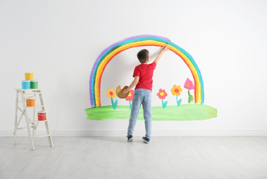 Image of Little child drawing rainbow and flowers on white wall indoors