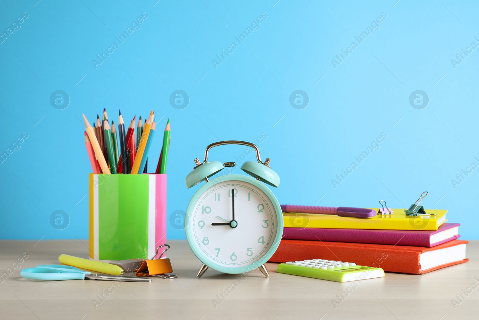 Photo of Turquoise alarm clock and different stationery on white wooden table against light blue background. School time