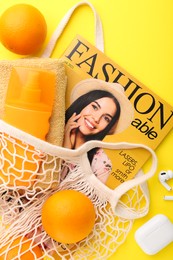 String bag with fresh oranges, fashion magazine and beach accessories on yellow background, flat lay