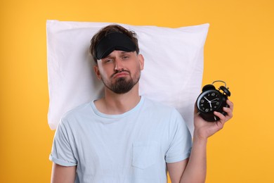 Photo of Unhappy man with pillow, alarm clock and sleep mask on yellow background. Insomnia problem