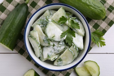 Delicious cucumber salad in bowl and ingredients on white wooden table, flat lay