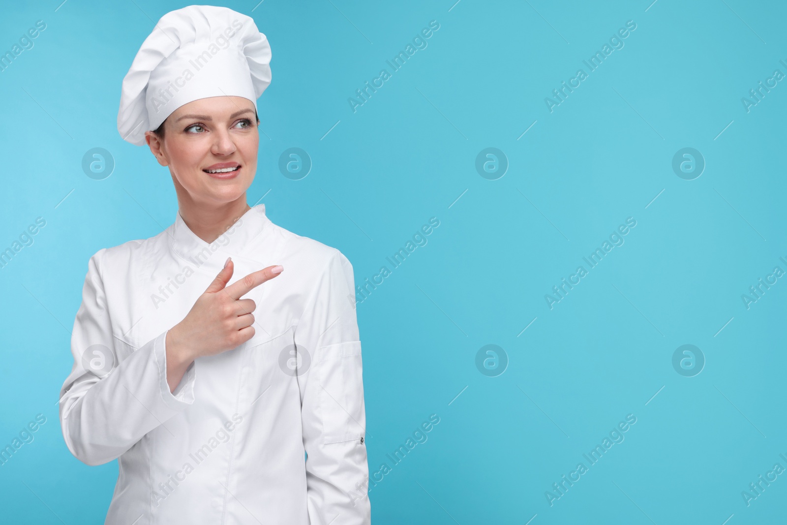 Photo of Happy woman chef in uniform pointing at something on light blue background, space for text