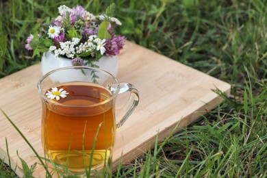 Cup of aromatic herbal tea and ceramic mortar with different wildflowers on green grass outdoors. Space for text