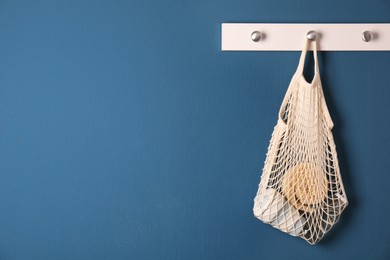 Photo of Conscious consumption. Net bag with eco friendly products hanging on blue wall indoors, space for text