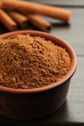 Bowl of cinnamon powder and sticks on wooden table, closeup