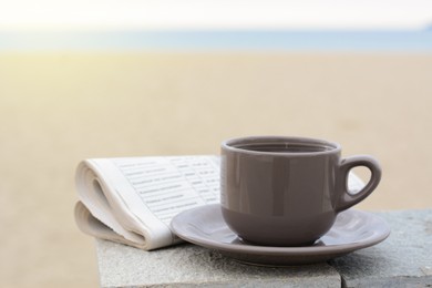 Ceramic cup of hot drink and newspaper on stone surface near sea in morning