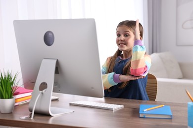 Photo of E-learning. Cute girl raising her hand to answer during online lesson at table indoors