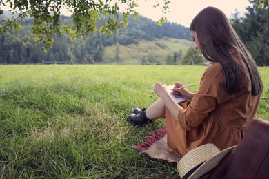 Photo of Beautiful young woman drawing with pencil in notepad outdoors on green grass