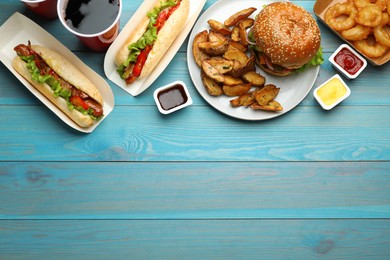 Burger, potato wedges, fried onion rings, hot dogs and refreshing drink on light blue wooden table, flat lay with space for text. Fast food