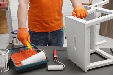 Photo of Man taking orange paint with roller from tray at grey wooden table indoors, closeup