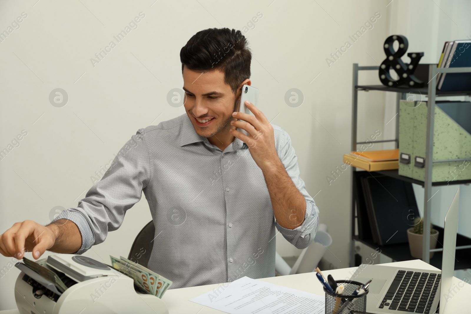 Photo of Man using banknote counter while talking on phone at white table indoors