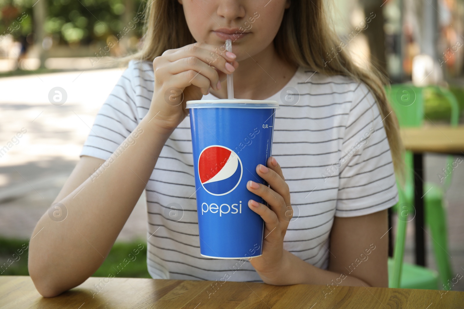 Photo of MYKOLAIV, UKRAINE - JUNE 9, 2021: Woman drinking Pepsi from paper cup at table in outdoor cafe, closeup
