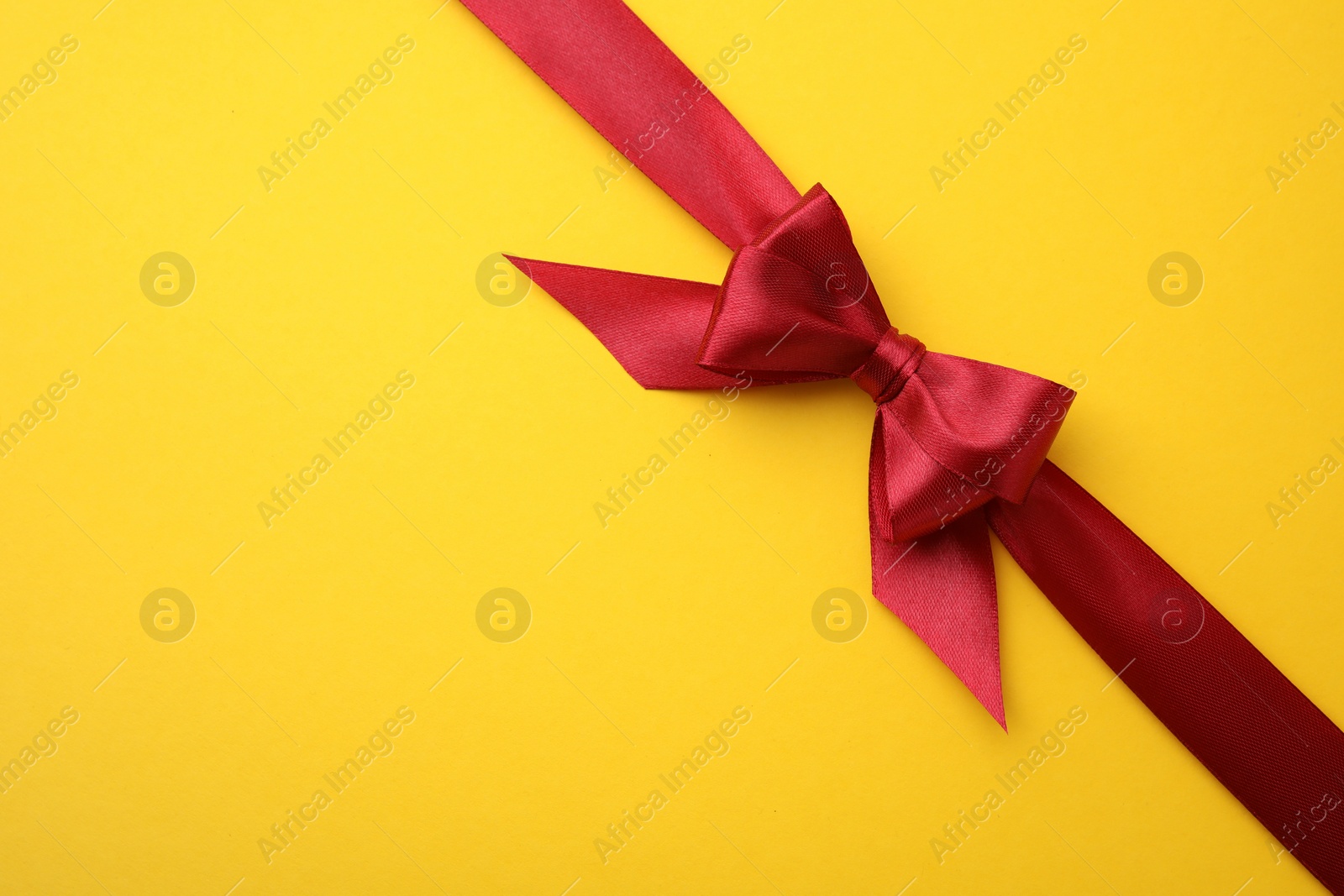 Photo of Red satin ribbon with bow on yellow background, top view. Space for text