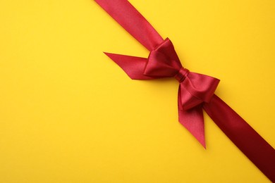 Photo of Red satin ribbon with bow on yellow background, top view. Space for text