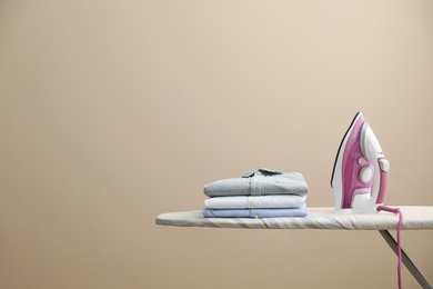 Photo of New modern iron and clean shirts on board against beige background, space for text. Laundry day