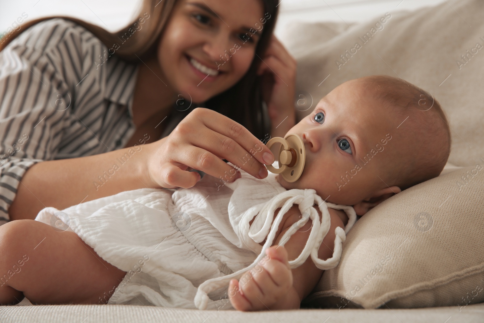 Photo of Happy mother giving pacifier to her cute little baby on sofa at home