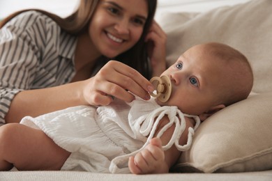 Happy mother giving pacifier to her cute little baby on sofa at home