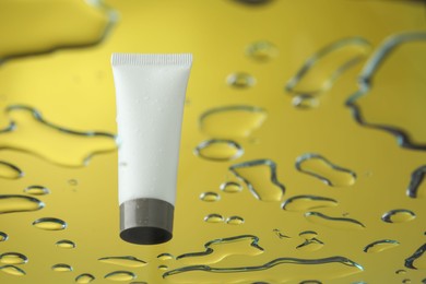 Photo of Moisturizing cream in tube on glass with water drops against yellow background, low angle view. Space for text