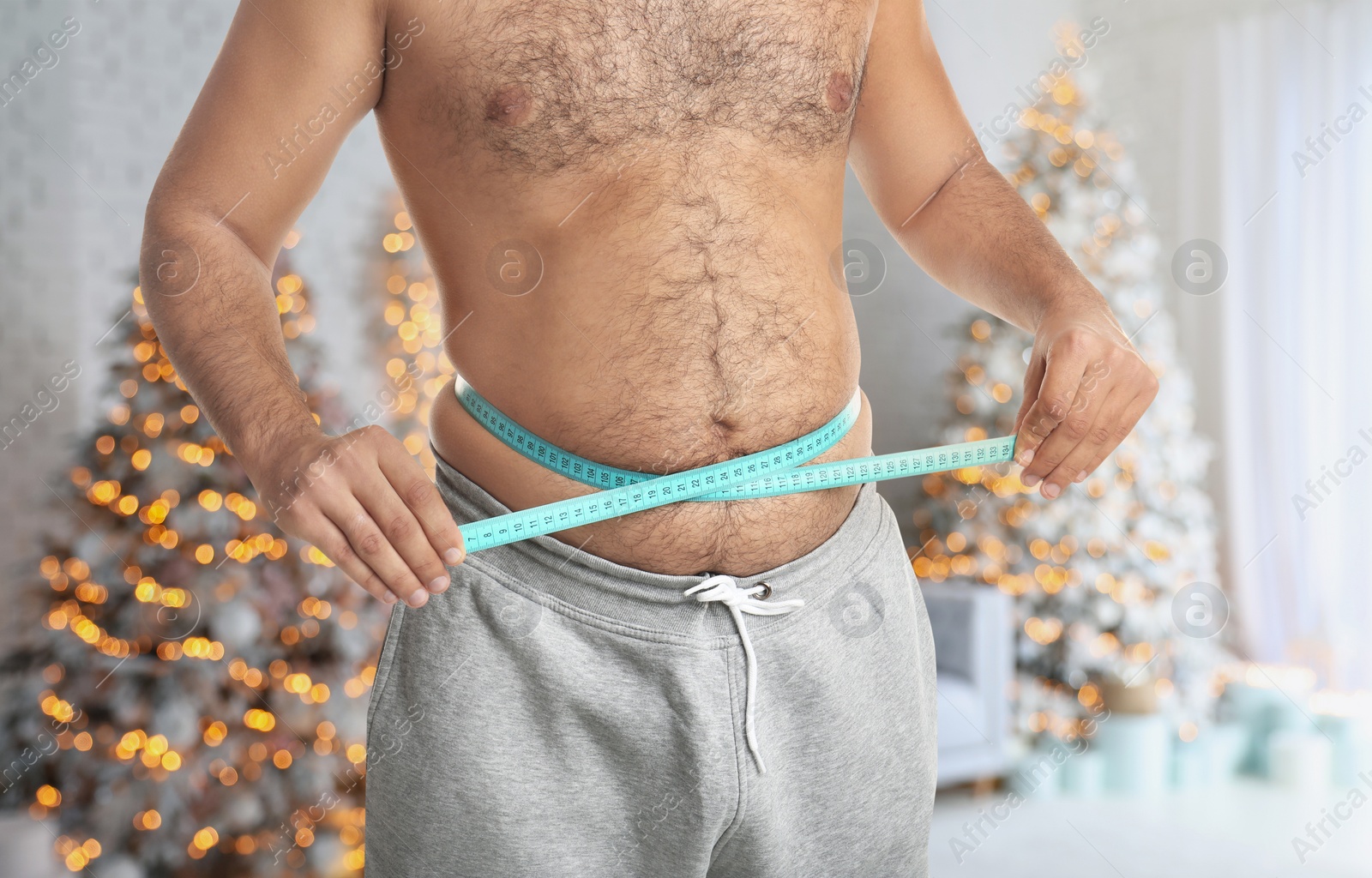 Image of Overweight man measuring his waist in room with Christmas trees after holidays, closeup