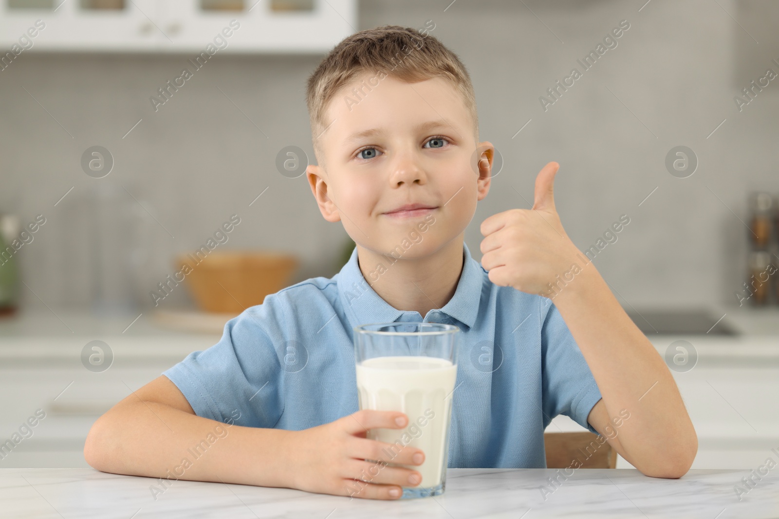 Photo of Cute boy with glass of fresh milk showing thumb up at white table in kitchen