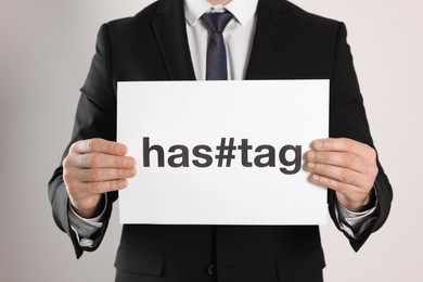Photo of Man holding sheet of paper with word and symbol Hashtag on light background, closeup