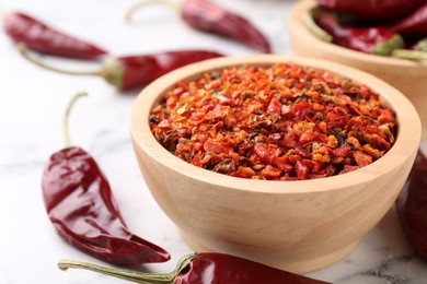 Photo of Chili pepper flakes in bowl and pods on white marble table, closeup