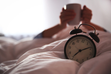 Woman with cup in bed, focus on alarm clock. Morning time
