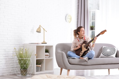 Young woman with headphones playing electric guitar in living room. Space for text