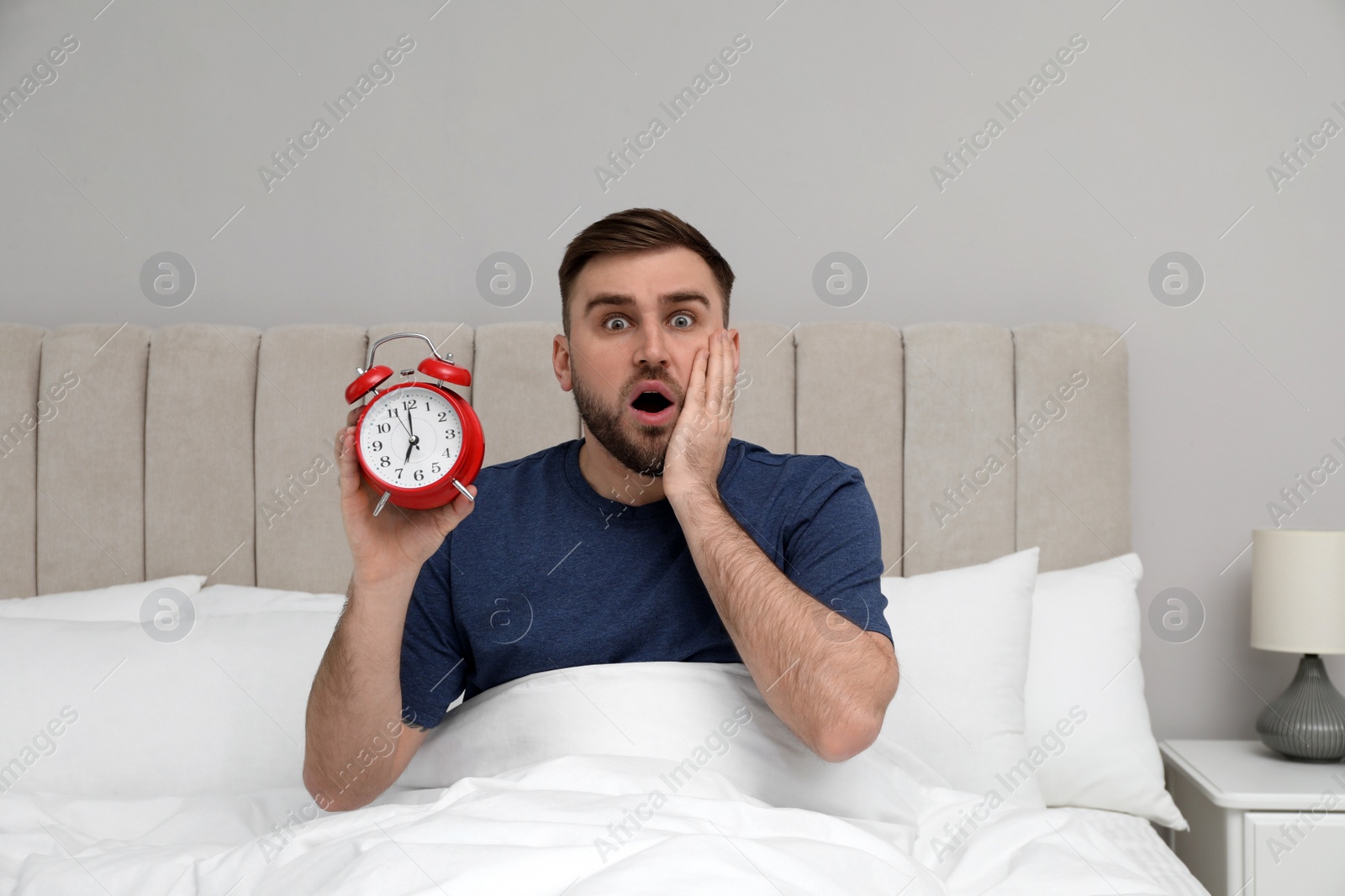 Photo of Emotional overslept man with alarm clock in bed. Being late concept