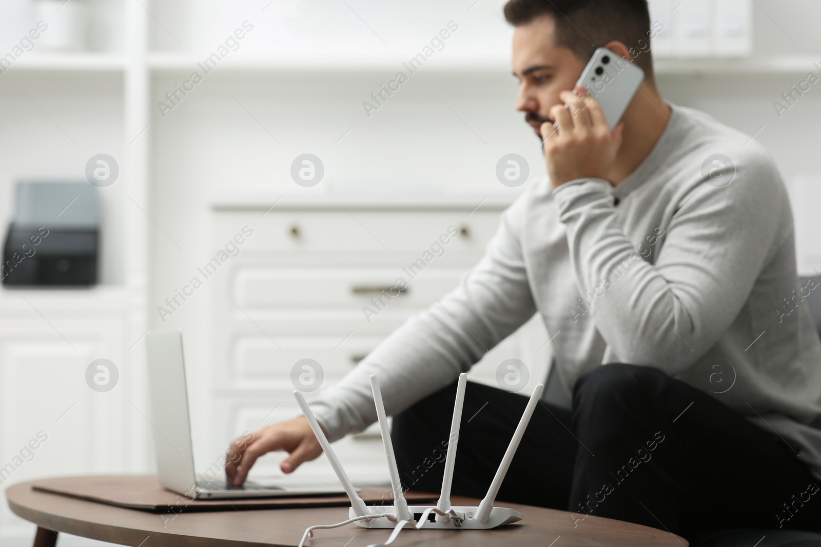 Photo of Man talking on smartphone while working at wooden table indoors, focus on Wi-Fi router