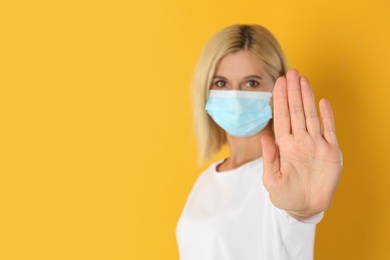 Photo of Woman in protective mask showing stop gesture on yellow background, space for text. Prevent spreading of coronavirus