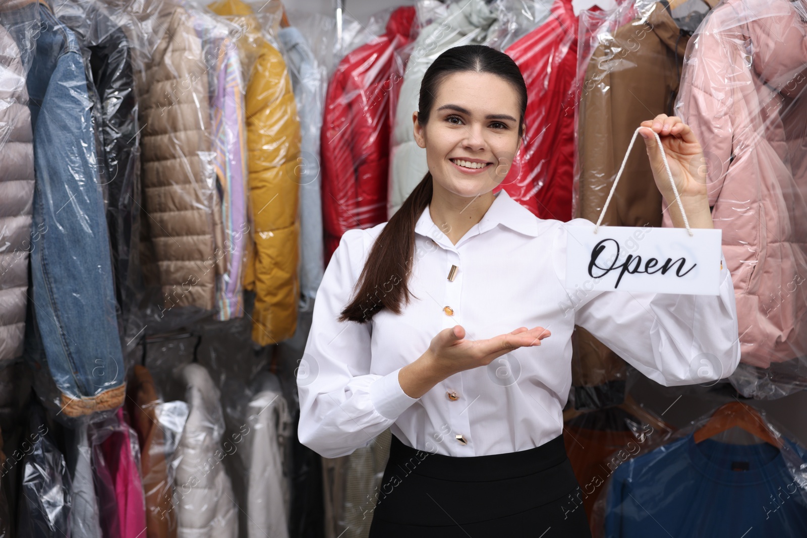 Photo of Dry-cleaning service. Happy worker holding Open sign near rack with clothes indoors, space for text