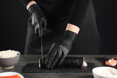 Chef in gloves cutting sushi roll at dark textured table, closeup