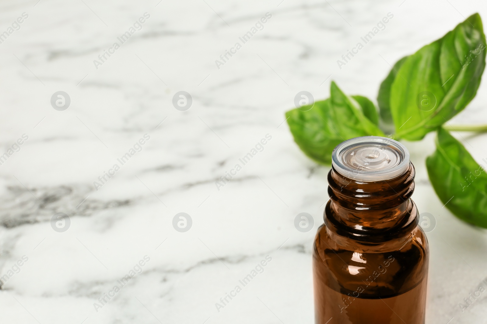 Photo of Bottle of basil essential oil and fresh leaves on marble table. Space for text
