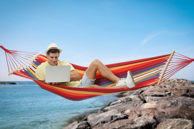 Man with laptop resting in hammock near sea on sunny day 