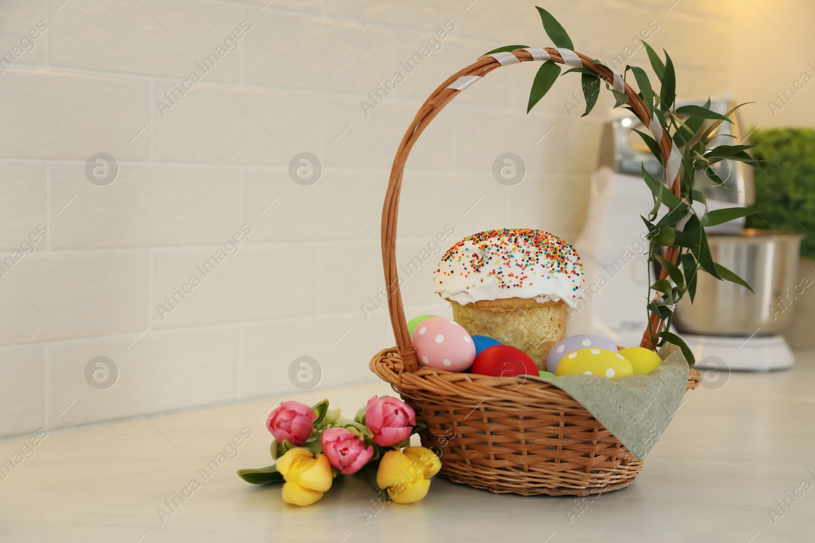 Photo of Basket with traditional Easter cake, dyed eggs and flowers on table near white brick wall. Space for text