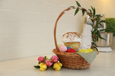 Basket with traditional Easter cake, dyed eggs and flowers on table near white brick wall. Space for text