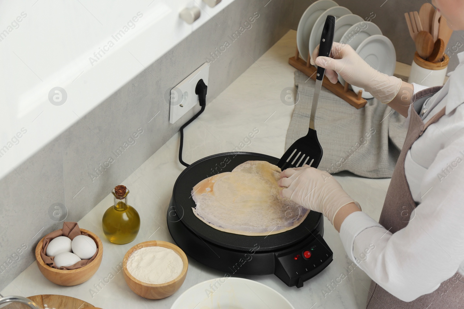 Photo of Woman cooking delicious crepe on electric pancake maker at white marble table in kitchen, closeup